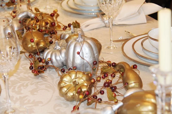 berries in yellow and dark red, and gold and silver, spray-painted pumpkins and gourds, decorating a pale cream ornamental tablecloth, with glasses and gold-rimmed stacked plates