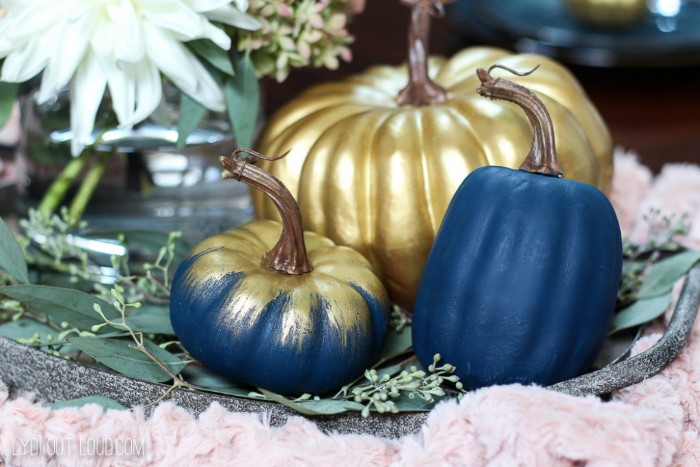 three pumpkins painted in dark blue, and metallic gold paint, thanksgiving card messages, white flowers and green plants in the background