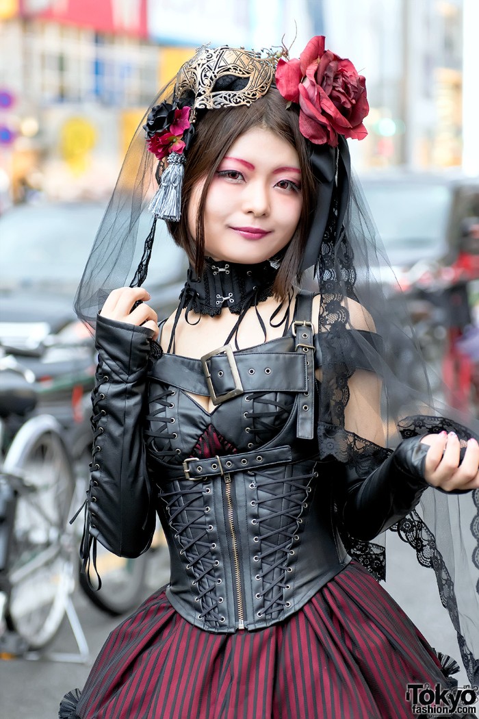leather corset in black, with several belts, worn over a striped dress, in purple and black, by a smiling young gothic lolita, with faux flowers, and a venetian mask in her hair