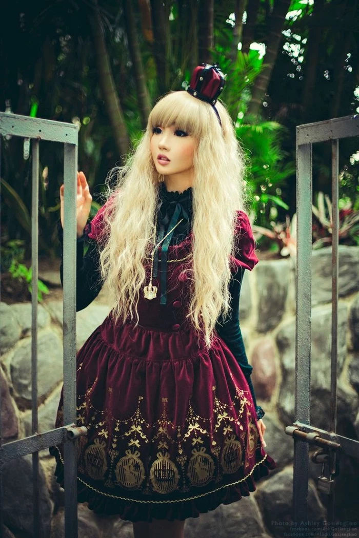 velvet dress in burgundy, with gold print, worn over a black lacy shirt, by a japanese lolita, in a long blonde wig, decorated with a miniature crown