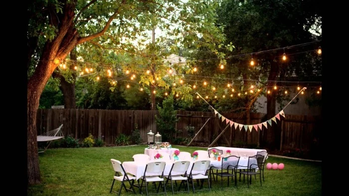 trees decorated with string lights, and a colorful banner, near two tables, set up for a festive meal, 50th birthday party ideas for mom, pink balloons in a garden