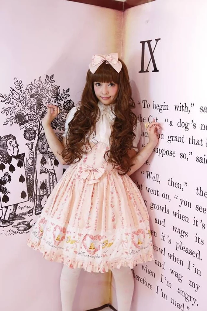 giant book behind a girl, in a long curled brunette wig, with straight bangs, wearing a light peach pink and white dress, with cutesy print, frills and bows, sweet lolita