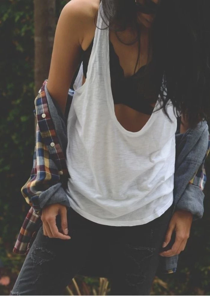 flannel shirt worn off the shoulders, by a slim woman, in a loose white tank top, over a black bralette, dark grey jeans