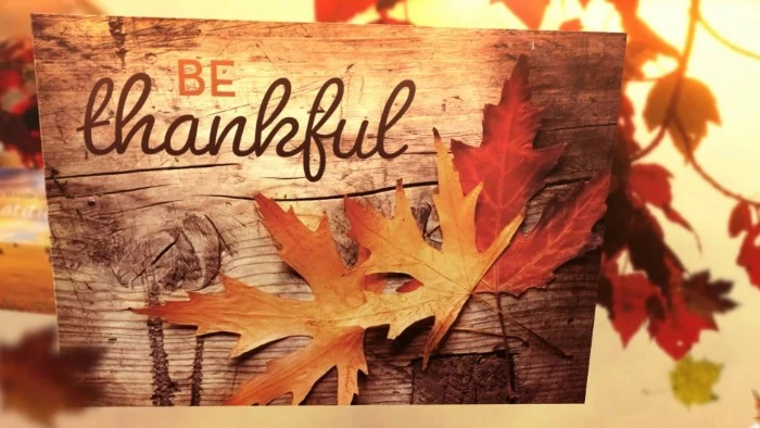 rectangular piece of wood, decorated with orange and red fall leaves, bearing the inscription be thankful, written in orange and dark brown