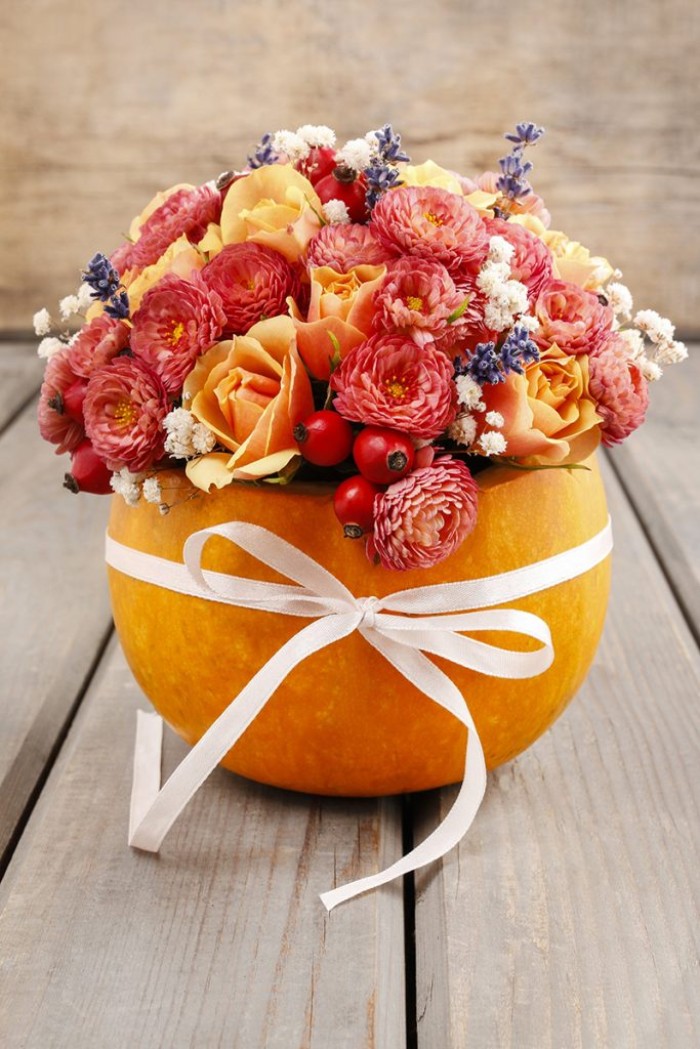 ribbon in white, tied in a bow around a large, hollow orange pumpkin, filled with red flowers, pale orange roses, lavender and small white blossoms, cheap centerpiece ideas, on a pale grey wooden surface