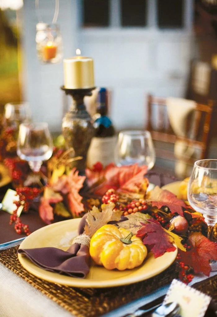burgundy red and orange, yellow and beige fall leaves, on a table with wine glasses, a gold-colored lit candle, and a yellow plate, containing a small yellow pumpkin