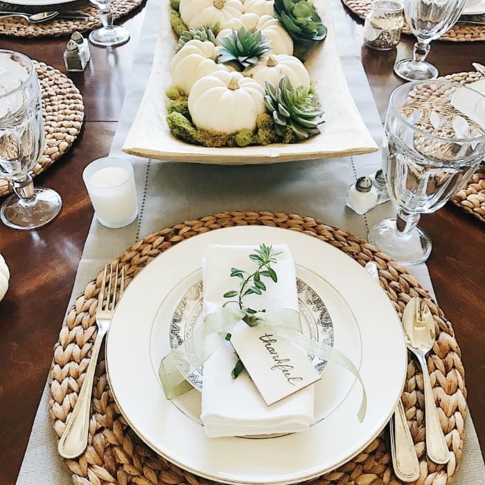 sprig of a green plant, tied to a folded white napkin, with a sheer silber ribbon, featuring a small label saying thankful, placed on a plate, thanksgiving table setting, round rattan table mats