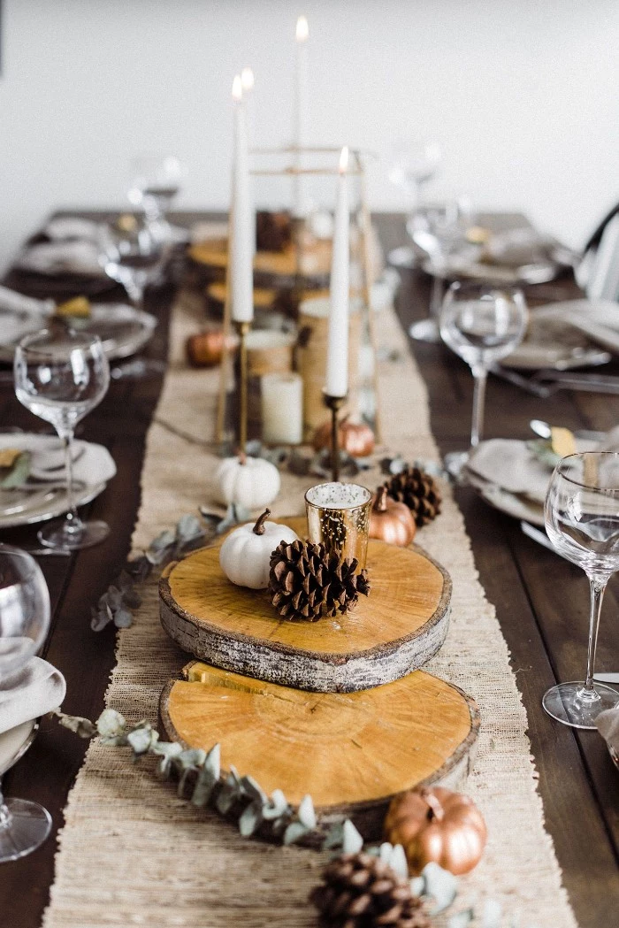 rectangular table set up for a festive meal, thanksgiving greetings, center piece featuring natural wood pieces, pine cones and small decorative pumpkins, tall lit candles