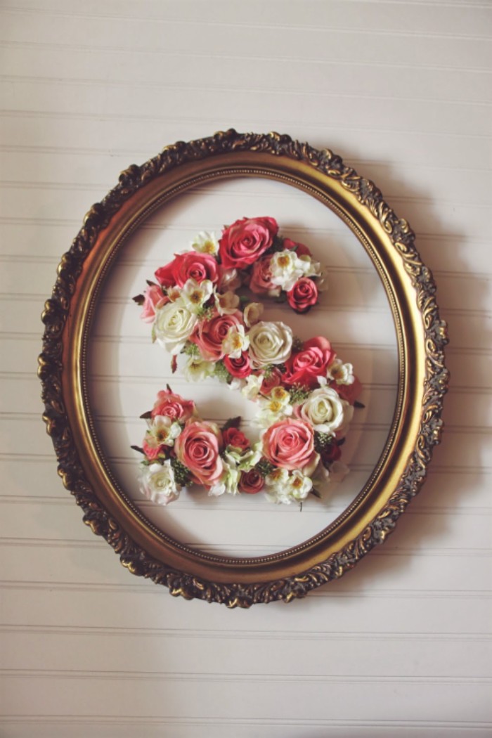 oval frame in gold, with ornamental details, containing the letter s, decorated with faux flowers, pink and white roses, white and yellow blossoms