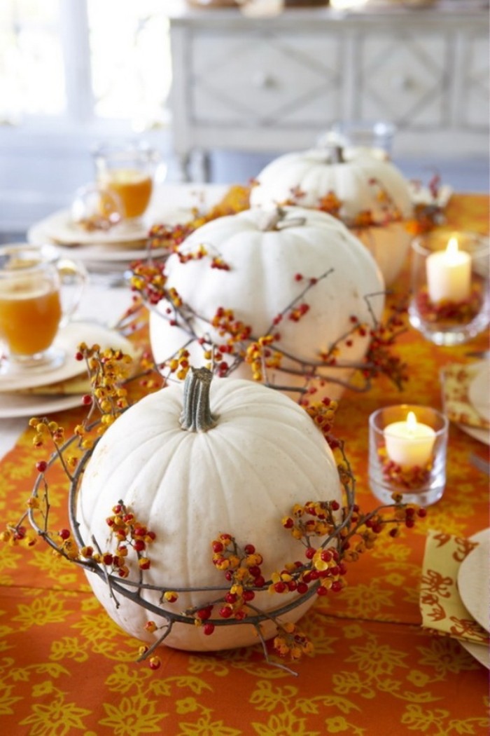 wreaths made from thin branches, with tiny orange berries, wrapped around three white pumpkins, placed on an orange and yellow patterned tablecloth