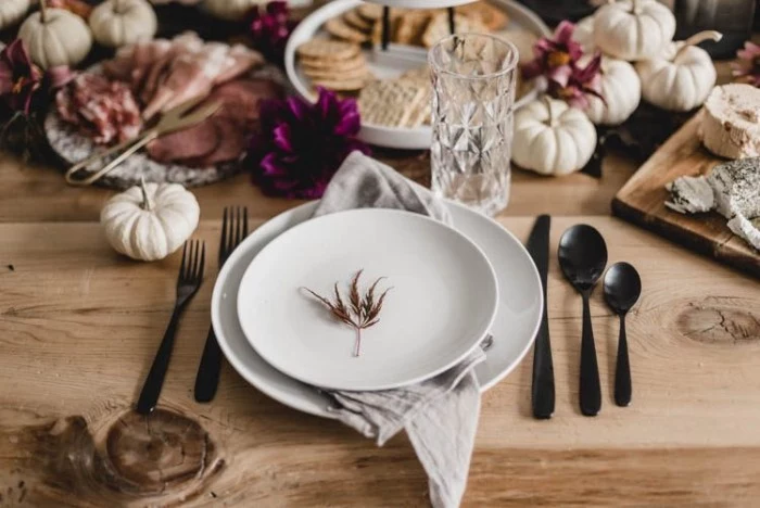 little fall leaf, placed in the middle of a round white plate, surrounded by black cutlery, on a wooden table, with various foods, and small white decorative pumpkins, thanksgiving table setting