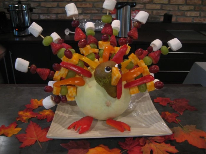 marshmellow and fruit kaboobs, pinned on a pale pumpkin, made to resemble a turkey, with fruit and vegetables, turkey decorations, on a large white square plate, surrounded by fall leaves 