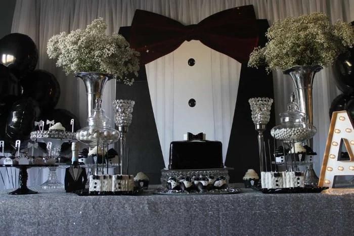 wall decoration shaped like a large tuxedo, near a table with a black cake, various black and white sweets, and two bouquets of small white flowers
