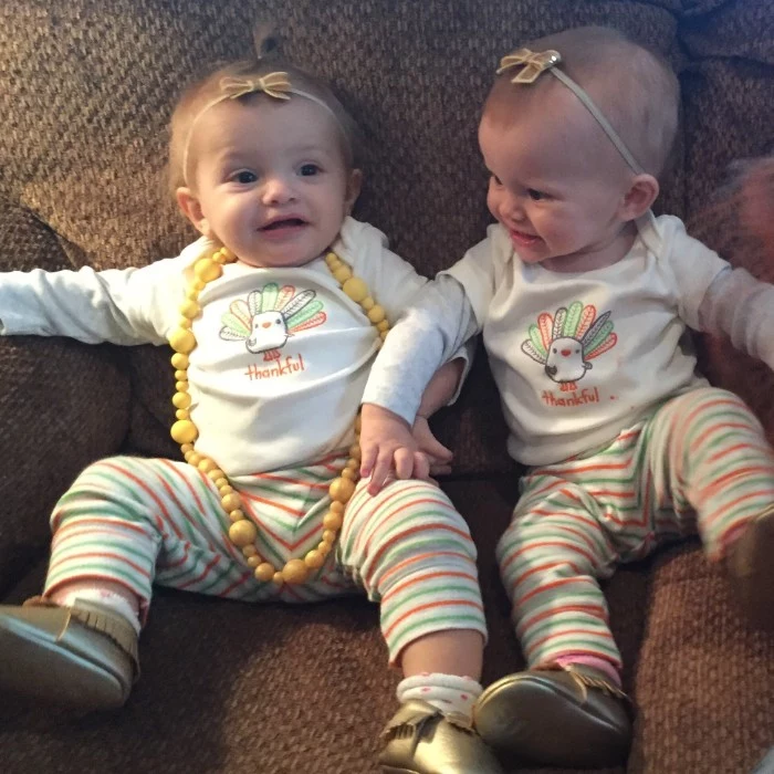 pair of babies, dressed in identical clothes, white jumpers featuring cartoon turkey prints, baby girl thanksgiving outfit, striped trousers in light green, white and orange, shiny gold soft shoes