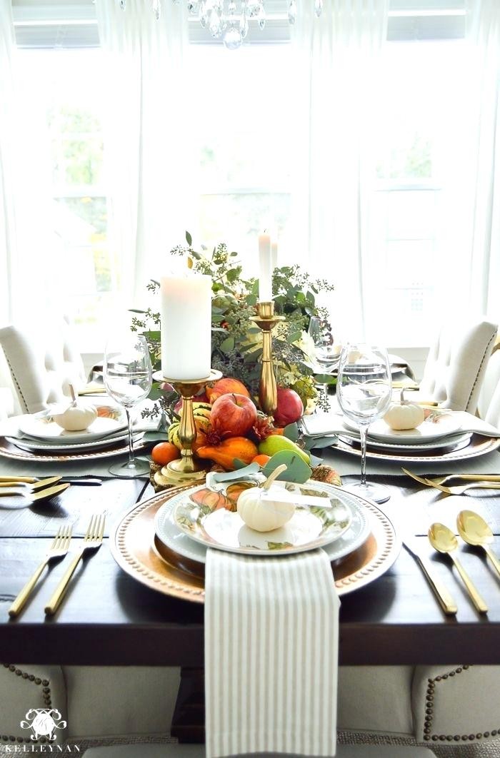 well lit room with a black table, set for a festive meal, stacked plates, decorated with small white pumpkins, gold cutlery and striped napkins, thanksgiving dinnerware, candles and flowers, fruit and gourds
