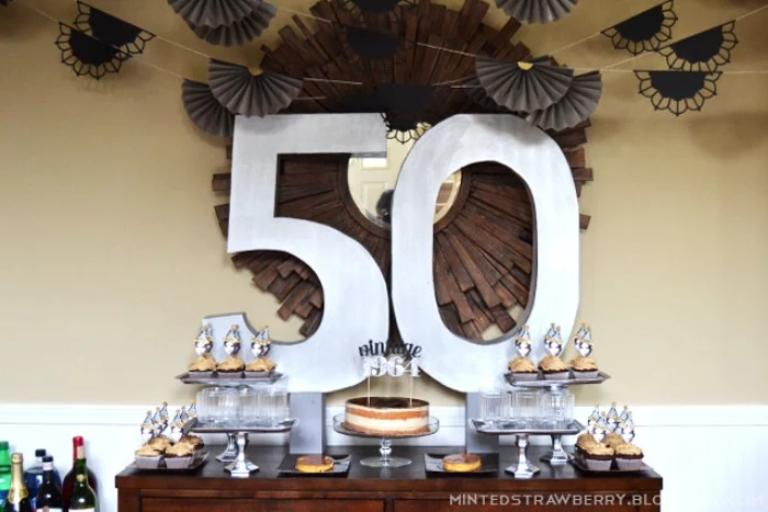 big wall decoration, featuring the numbers 5 and 0, surrounded by grey, fan-like paper garlands, near a table with a large cake, and some cupcakes, 50th birthday celebration ideas for husband