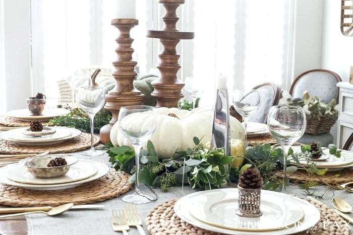 round knitted rattan table mats, white plates and wine glasses, on a table, decorated with wooden candleholders, green leaves and pinecones, thanksgiving table decorations, large white pumpkin