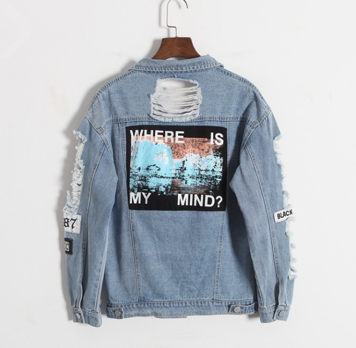 denim jacket in light blue, with several large rips, and a few patches, featuring a graphic print on its back, on a wooden hanger