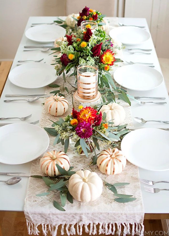 purple and red, yellow and orange flowers, with green leaves, in three bouquets, surrounded by small white pumpkins, some decorated with gold stripes, thanksgiving centerpiece, placed on a white rectangular table