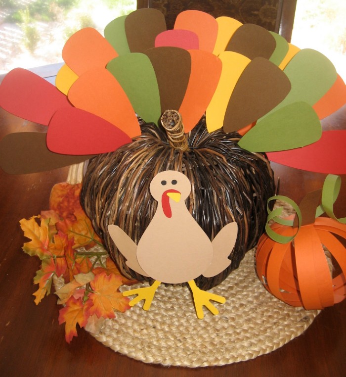paper cutout of a turkey, stuck on a whicker pumpkin ornament, with a tail made from pieces of paper in different colors, turkey decorations, faux fall leaves, and a paper pumpkin nearby