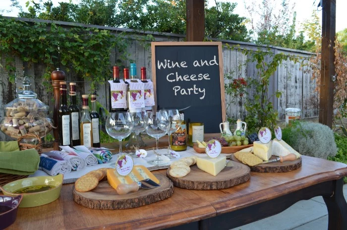 table with several cheese platters, wine bottles and glasses, small blackboard featuring the inscription, wine and cheese party, 50th birthday party ideas