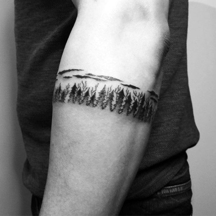 cool arm tattoos, a bracelet-like black tattoo, going around a man's arm, just below his elbow, featuring multiple trees and clouds
