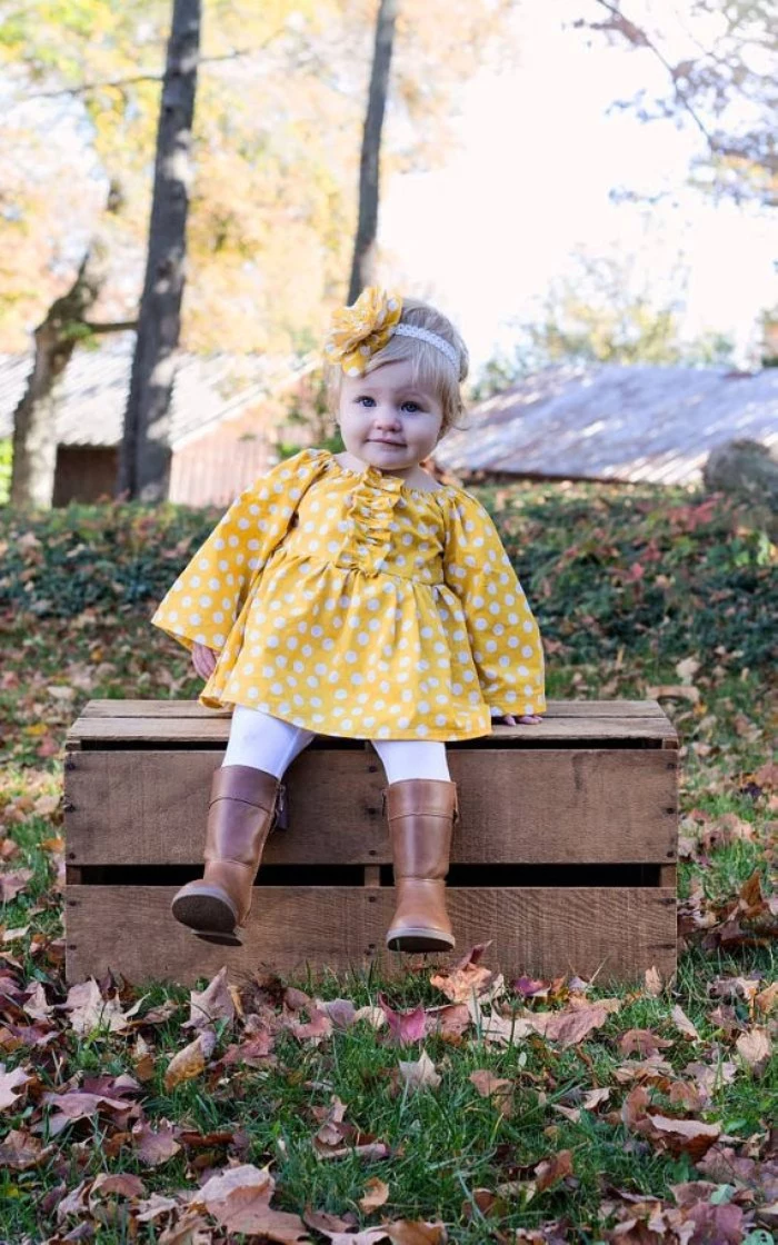 polka dots in white, on a yellow dress, with long sleeves and frills, worn by a smiling baby girl, with brown leather boots, white leggings and a yellow and white head ornaments