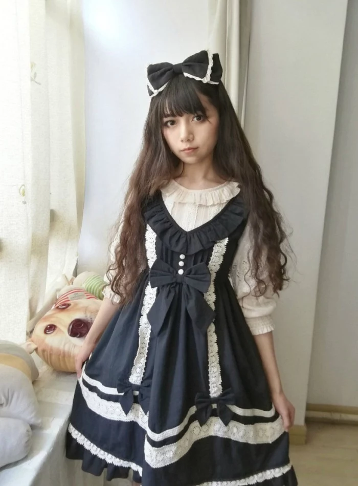 v-neck pinafore dress, in dark navy blue, featuring white lace and decorative bows, worn over a frilly white blouse, by a japanese lolita, with long dark brunette hair
