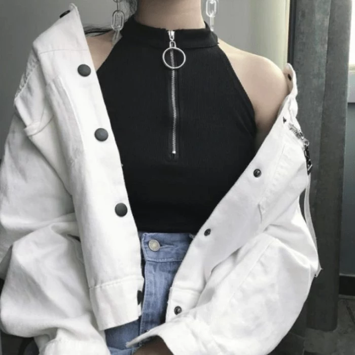 white baggy jacket, with black click-clack buttons, worn over a sleeveles black top, with a high collar, and a zip-up detail, pale blue jeans, and chain-like earrings