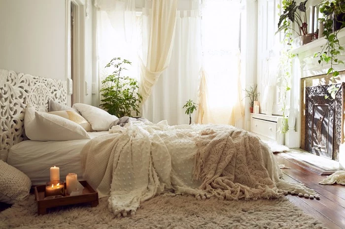 double bed with white, cream and light beige pillows, linen and blankets, in a room with several green potted plants, a fireplace and a window with light curtains