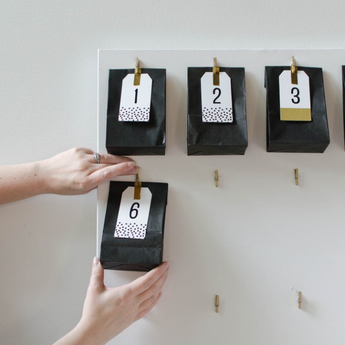 pair of hands, attaching a black paper bag, with a white label, featuring the number 6, onto a white board, fun advent calendars, more black paper bags attached above