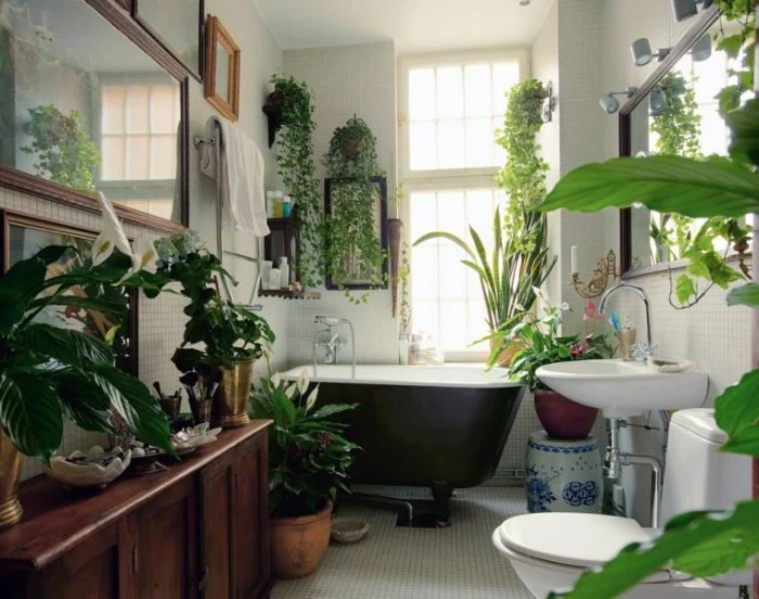 lots of green potted plants, in a boho style bathroom, with a black and white tub, a wooden cupboard, a sink and a toilet, and several framed images