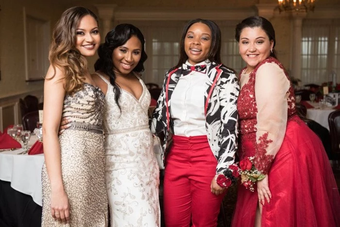friends smiling at the camera, wearing smart outfits, beaded embroidered gowns, in pale beige and white, a red dress with lace, and sheer white sleeves, cocktail attire for women, red trousers with a white shirt, and a black, white and red jacket, worn with a matching bowtie