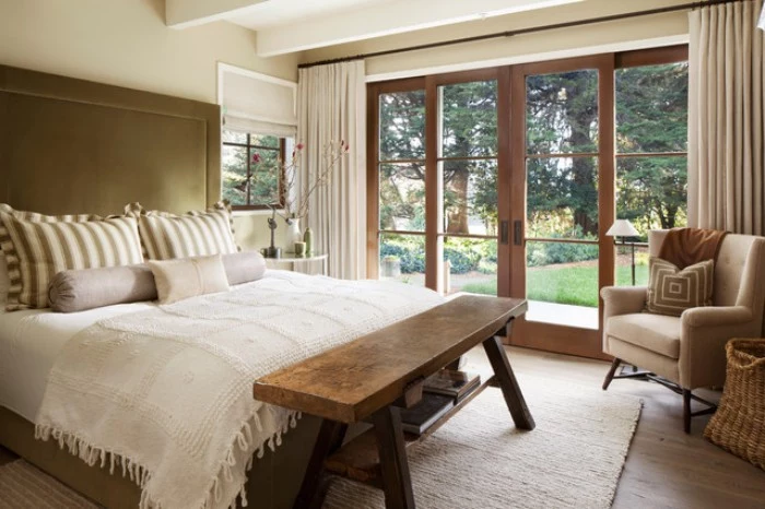 wooden bench and a pale beige armchair, in a bedroom with cream walls, a white ceiling, and a double bed, with a brown headboard