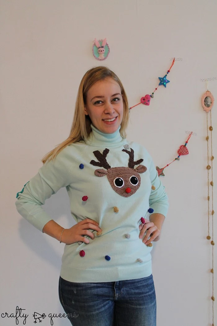 diy ugly christmas sweater, smiling blonde woman, dressed in a pale blue jumper, decorated with small pom poms, in different colors, and a brown crocheted deer's head, with a red nose