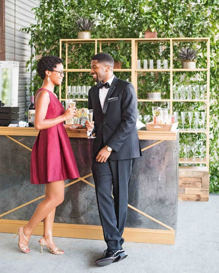 wine red mini dress, worn by a smiling woman, holding a drink and talking to a man, dressed in a smart black suit, cocktail attire for men, with a white shirt, and a black bowtie