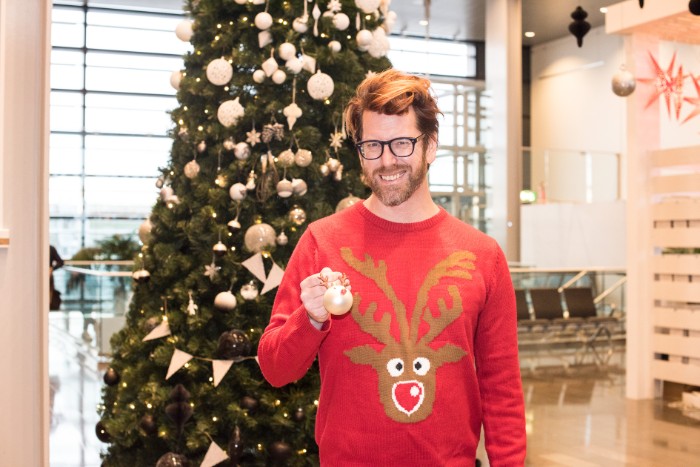 bespectacled man with a short beard, wearing a red sweater, featuring an image of a brown cartoon deer's head, with a red nose