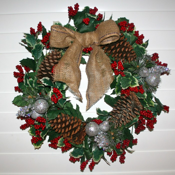 fir cones and small red berries, on a diy christmas wreath, made from green leaves, and decorated with small silver baubles, and a large burlap bow