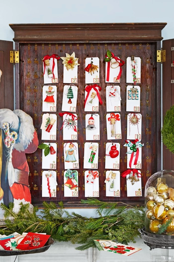 christmas countdown calendar, 25 small numbered white cards, each containing a small christmas tree decoration, placed inside a shallow, wooden cupboard-like structure, surrounded by festive decorations
