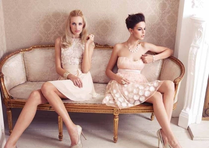light peach occasion mini dresses, with white lace details, cocktail attire for women, worn by two slim models, sitting on an antique pale beige couch