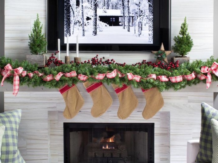 painting or photo of a snow covered lodge, framed above a fireplace, decorated with a green pine garland, featuring a red and white plaid ribbon, and four beige stockings