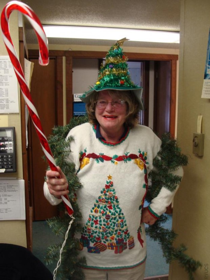 hat in green, shaped like a christmas tree, and decorated with garlands and baubles, worn by an elderly woman, dressed in a white jumper, featuring an xmas tree, and holding a giant peppermint cane