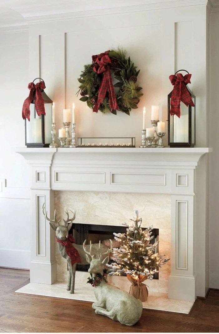 couple of deer statuettes, and a miniature christmas tree, decorated with glowing string lights, near a white and pale cream fireplace, images of christmas, green wreath with a red bow, and burning candles
