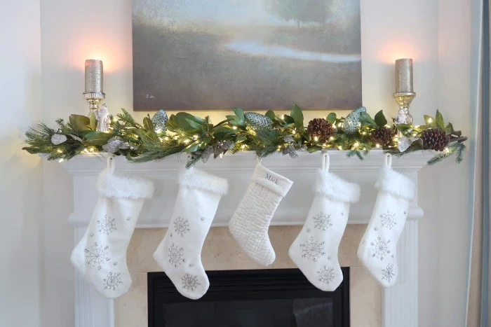embroidered white stockings, with silver snowflakes, hanging from a mantel, decorated with a garland, made from branches with large green leaves, and lit string lights, mantel decor for your home