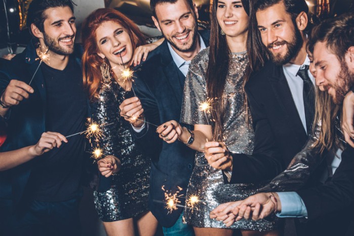 lit sparkles held by a group of smiling people, men wearing dark suits, some with pale shirts and black ties, women in sparkling silver sequin dresses, black tie optional outfit ideas