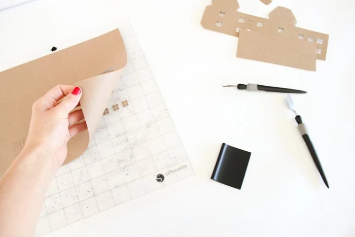 nail polish in red, on a hand, pulling a piece of beige card, away from a sheet of white, see through lined paper, adult advent calendar making process, two pen knives, and some cutouts nearby