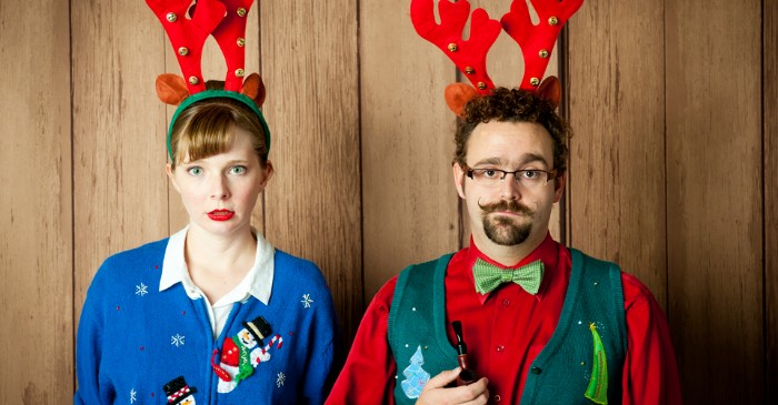 couple with red and green headbands, featuring deer ears and antlers, the woman is wearing a blue cardigan with snowmen, and the man is dressed in a red shirt, forest green vest, and light green bowtie, ugly christmas sweater ideas for grownups