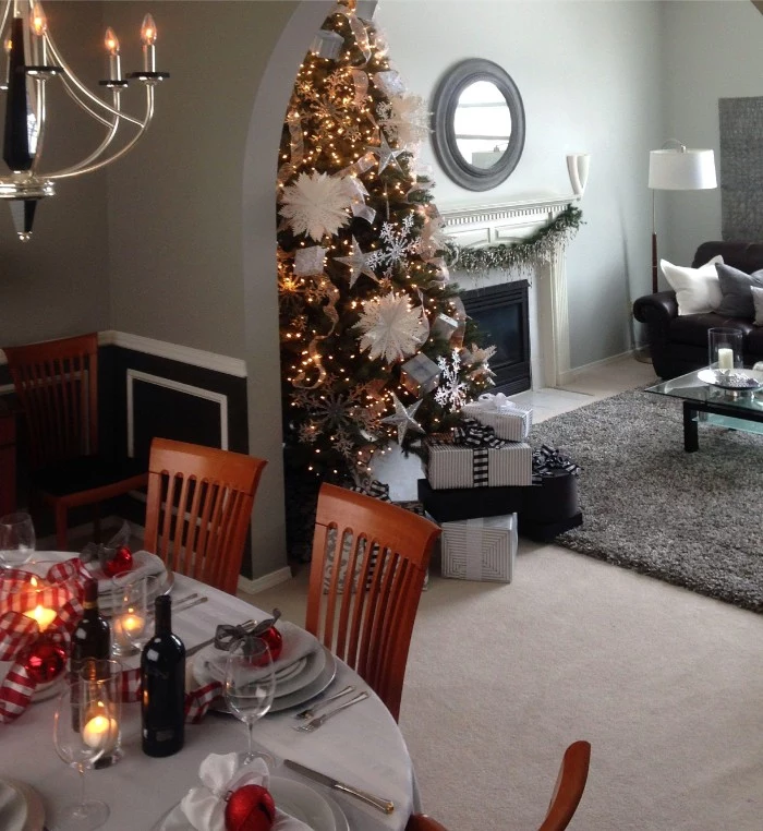 table set for a festive dinner, in a dining area, overlooking a living room, decorated with a christmas tree, covered in white ornaments, and glowing string lights, holiday images, presents and a festive garland