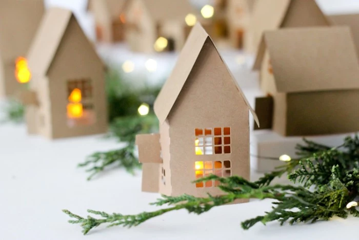adult advent calendar tutorial, small cardboard houses, with cutout windows and doors, each containing a lit tea light, forming a christmas village, surrounded by pine branches