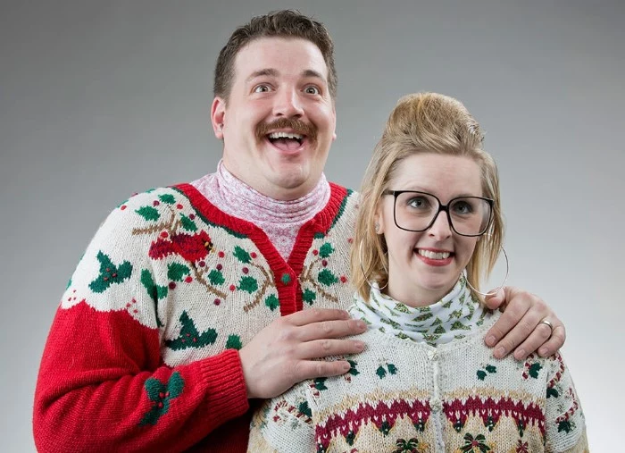 cardigan in red and white, with green holly branches, and a robin, worn by a smiling man, with a mustache, and a pale pink turtleneck, hugging a blonde woman,in a similar kitsch retro cardigan